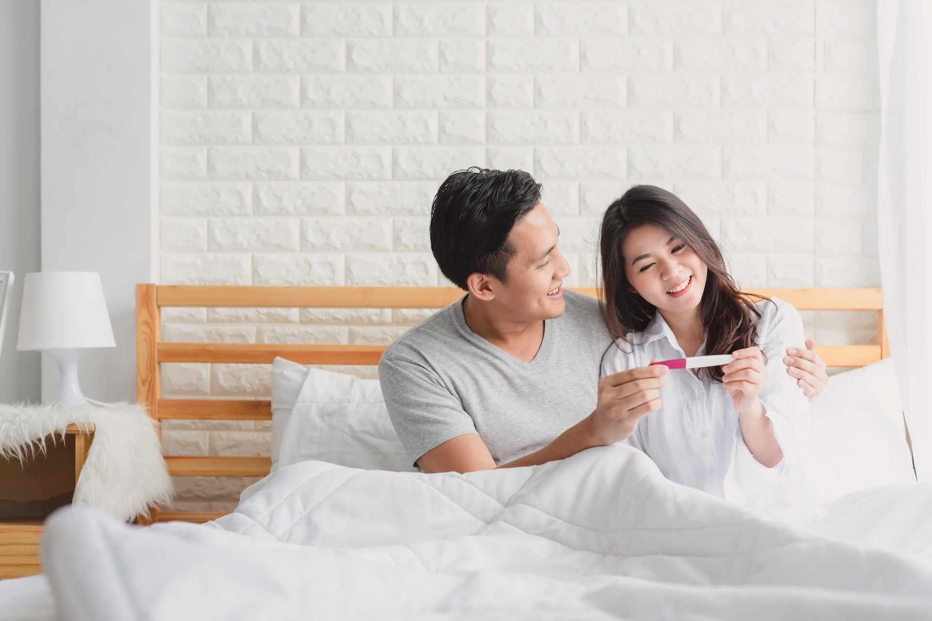 Asian couple smiling at their pregnancy test result in bed