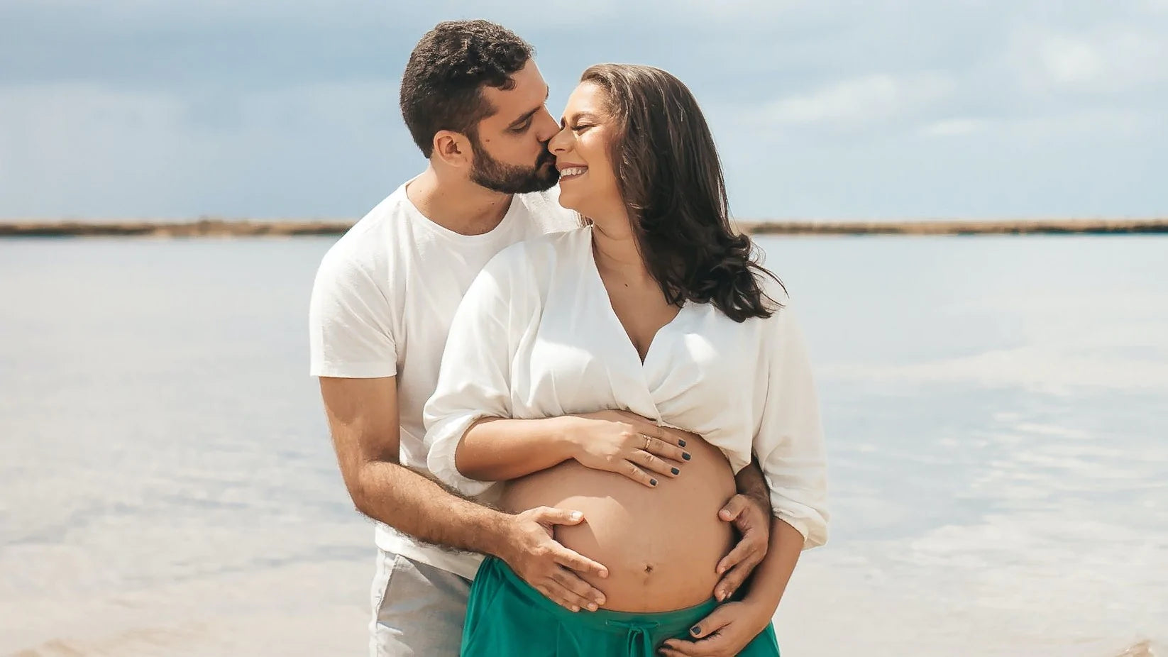 husband kisses and embraces pregnant wife on the beach